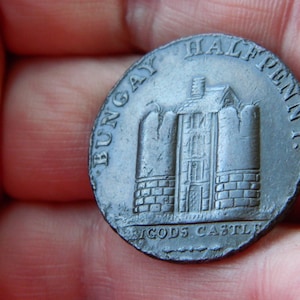 Peut inclure: Une pièce de monnaie ancienne avec l'inscription "BUNGAY HALF PENNY" et "GODS CASTLE" entourant une représentation détaillée d'un bâtiment. La pièce, tenue dans une main, montre des signes d'usure et une couleur métallique.
