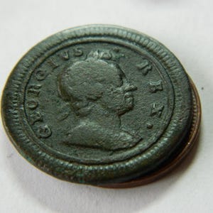 Könnte beinhalten: Nahaufnahme einer dunkelgrünen antiken Münze. Die Münze zeigt das Profil einer Person und die Inschrift "GEORGIVS REX". Die Münze ist leicht abgenutzt und weist Gebrauchsspuren auf. Die Münze liegt auf einer anderen Münze.