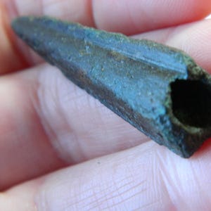 May include: A weathered, bronze-coloured artifact with a hollow, rectangular opening at one end. The object has a tapered shape and a rough, textured surface, suggesting age and exposure. The colour is a mix of bronze and green.