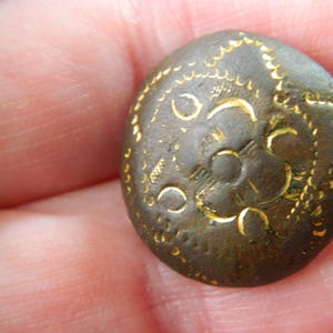 Puede incluir: Un botón redondo de bronce antiguo con diseños florales y de media luna grabados en color dorado. El botón tiene una forma ligeramente abovedada y está sostenido en la mano de una persona. La superficie del botón tiene un aspecto texturizado.