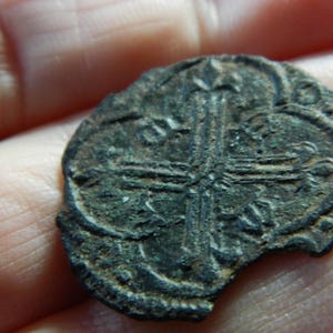 Puede incluir: Una moneda oscura y desgastada con un diseño de cruz en el centro. La moneda tiene forma ovalada con un borde roto, y parece estar hecha de un metal oscuro. La superficie muestra signos de edad y desgaste.