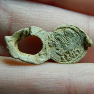 Puede incluir: Un pequeño objeto metálico desgastado con una abertura circular y un diseño circular con bisagras. El objeto tiene un diseño en relieve en un lado y el número "1840" es visible. El objeto es de un color gris claro apagado.