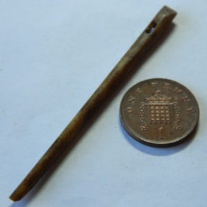 May include: A brown, slender, wooden tool with a hole near the top, possibly a needle or awl, lies next to a British one penny coin. The coin is bronze and shows a crowned emblem. The tool is approximately 5 inches long.