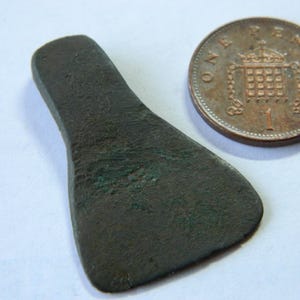 May include: A small, dark bronze artifact with a rounded blade and a handle-like extension. A copper penny is next to the artifact for size comparison. The artifact has a weathered appearance, suggesting age.