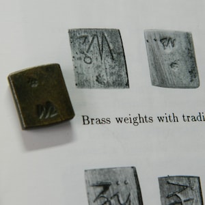 Könnte beinhalten: Nahaufnahme von Messinggewichten mit traditionellen Symbolen. Die Gewichte sind rechteckig mit abgerundeten Kanten und einem dunklen, gealterten Aussehen. Der Text "Brass weights with traditional symbols" ist sichtbar.