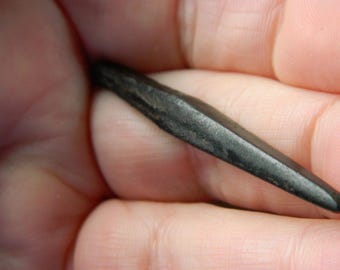 May include: A small, elongated, dark grey stone object with a tapered, pointed end and a slightly textured, matte surface. The object has a slender, needle-like shape with a thicker centre that gradually narrows towards the tip, showing natural variations.