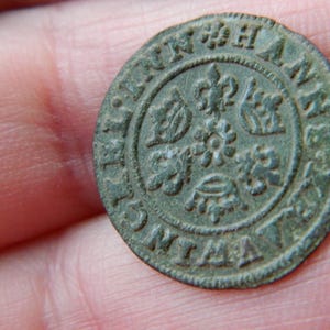 Puede incluir: Una moneda circular y desgastada con detalles en relieve. La moneda presenta motivos florales y coronas, junto con texto. La moneda tiene una pátina verde-marrón, lo que sugiere antigüedad. La moneda se sostiene en una mano.