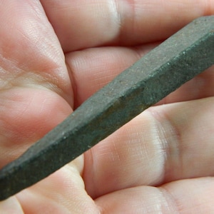 May include: A dark grey, wedge-shaped metal tool with a pointed end. The tool has a rectangular cross-section and appears to be made of a solid, heavy material. The tool is held in a person's hand.