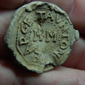 Puede incluir: Un artefacto circular y desgastado con letras en relieve. El texto incluye las letras "TA", "MM" y "NO", dispuestas alrededor del perímetro. El objeto tiene una superficie rugosa y texturizada y parece estar hecho de un material de color claro.