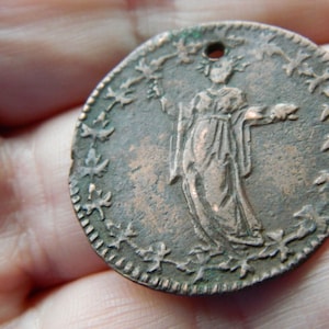 Puede incluir: Una moneda redonda de bronce antiguo que presenta una figura clásica con un vestido fluido, rodeada por una corona de hojas y estrellas. La moneda tiene un pequeño agujero en la parte superior y muestra signos de edad y desgaste.