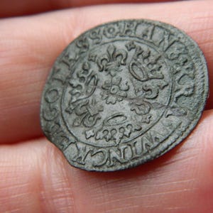 Puede incluir: Moneda antigua, redonda y deslustrada, con diseños florales y heráldicos en relieve. La superficie de la moneda muestra signos de desgaste y envejecimiento, con texto visible en los bordes. La moneda se sostiene en una mano.