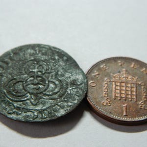Puede incluir: Dos monedas antiguas. Una es una moneda grande, oscura y redonda con un diseño ornamentado. La otra es una moneda más pequeña, de color cobre, con el texto "ONE PENNY" y una corona.