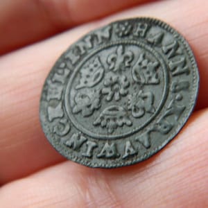 Puede incluir: Una moneda antigua y oscura con diseños florales y heráldicos en relieve. La superficie de la moneda muestra signos de desgaste, con texto visible en los bordes. La moneda se sostiene en una mano.