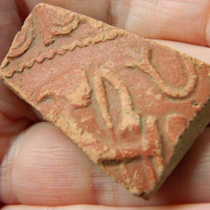 May include: A small, rectangular terracotta-colored clay fragment featuring an embossed, raised pattern of curved lines and geometric shapes. The surface has a matte, earthy texture with visible indentations and a worn, weathered appearance.
