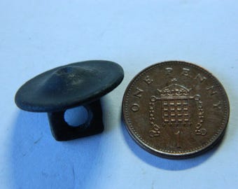 May include: A dark, matte-finished button with a slightly conical top and a square-shaped shank featuring a circular hole, positioned next to a copper-colored British one-penny coin for scale, providing a clear visual reference for its small size.