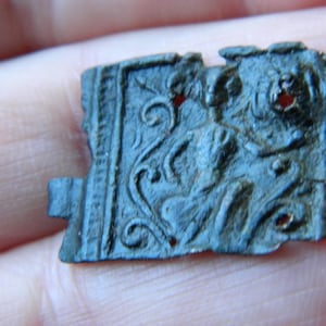 May include: A small, weathered, rectangular metal artifact with intricate relief carvings. The dark teal object features a central figure and swirling patterns, with a small tab extending from one side. The edges are slightly damaged.