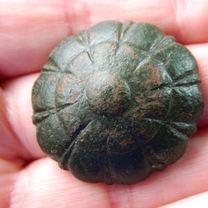 Peut inclure: Un bouton métallique circulaire vert foncé avec un motif en relief en forme de pétales de fleur rayonnant depuis un dôme central. La surface présente des signes d'oxydation et d'usure, avec des taches cuivrées visibles sous la patine verte.