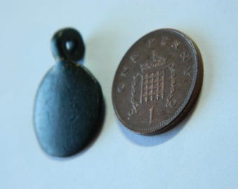 May include: A dark, oval-shaped pendant with a loop at the top, next to a one penny coin. The pendant is a dark, matte color. The coin is bronze-colored and shows the number 1 and a crown.