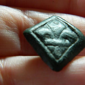 Può includere: Un bottone a forma di diamante verde scuro con un disegno a giglio in rilievo al centro. Il bottone ha una superficie leggermente strutturata e sembra essere in metallo. Il bottone è tenuto in una mano.