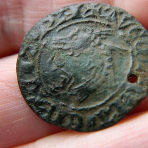Puede incluir: Una moneda circular de bronce desgastada, con un perfil en relieve de una persona coronada. La moneda tiene un pequeño agujero cerca del borde y está rodeada por un borde decorativo. La superficie de la moneda muestra signos de edad y desgaste.