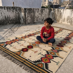 May include: A rectangular, cream-colored rug with a geometric pattern in shades of red, orange, green, and black. The rug has a fringed edge and a border with black and gold stripes. The rug is placed on a concrete surface.