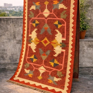 May include: A rectangular, rust-colored rug with a geometric pattern in cream, yellow, orange, and green. The rug has a fringed edge and a cream-colored border with a zigzag design. The rug is displayed outdoors.