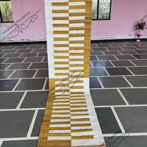 Puede incluir: Una alfombra rectangular con un patrón geométrico de rayas horizontales blancas y amarillo mostaza alternas. La alfombra está hecha de un material tejido y se muestra verticalmente.