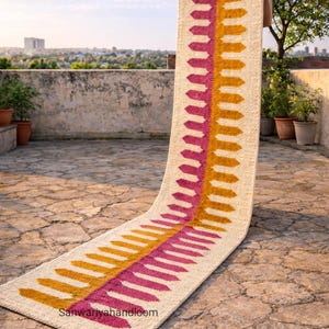 Puede incluir: Una alfombra rectangular larga con un fondo color crema y un diseño central de filas verticales de formas rosas y doradas en forma de flecha. La alfombra se exhibe al aire libre en un patio de piedra. El texto "Sanwariyahandloom" es visible.