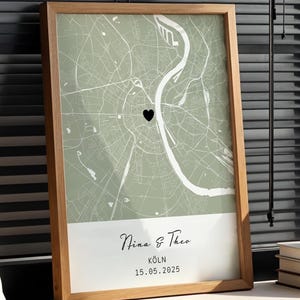 Puede incluir: Impresión enmarcada de un mapa de Colonia, Alemania, en un tono verde claro, con un corazón negro que marca una ubicación. La impresión incluye los nombres "Nina & Theo" y la fecha "15.05.2025". Una botella de vino y libros están al lado.