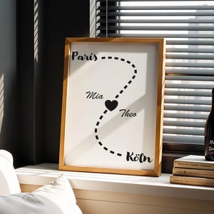May include: A framed print with a wooden frame, featuring a black dotted line connecting the cities of Paris, Mia, Theo, and Köln. A black heart is placed between Mia and Theo. A dark bottle of wine sits on a stack of books.