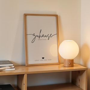 May include: A framed print with the word "zuhause" in script, a heart, and names. A round, white globe lamp with a wooden base sits on a wooden shelf with books. The scene is illuminated by the lamp.