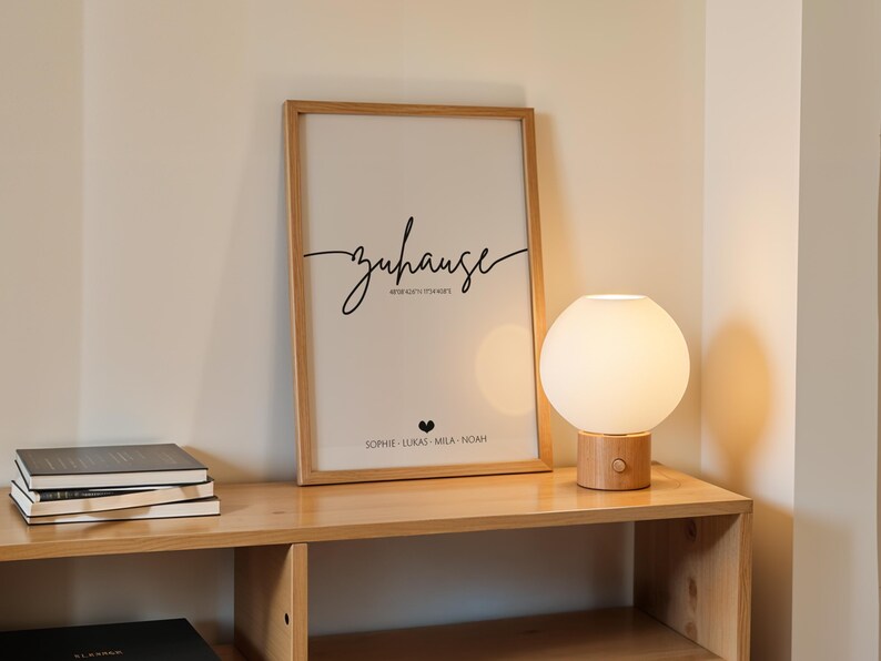 May include: A framed print with the word "zuhause" in script, a heart, and names. A round, white globe lamp with a wooden base sits on a wooden shelf with books. The scene is illuminated by the lamp.