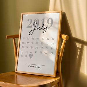 Può includere: Una stampa di calendario incorniciata per luglio 2019, esposta su una sedia di legno. Il calendario presenta un design minimalista con il mese e l'anno in alto e i giorni del mese in una griglia. I nomi "Elena & Theo" sono in basso.