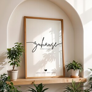May include: A framed art print with the word "zuhause" in cursive script, along with coordinates and names. The print has a light background and a wooden frame. Potted plants are arranged around the print.