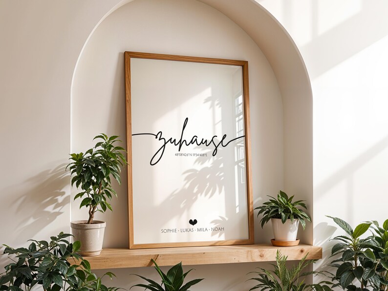 May include: A framed art print with the word "zuhause" in cursive script, along with coordinates and names. The print has a light background and a wooden frame. Potted plants are arranged around the print.