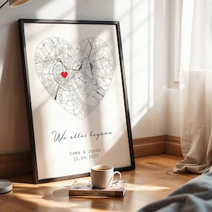 May include: Framed art print featuring a heart-shaped map with a red marker. The print includes the text "Wo alles begann", the names Emma & Jonas, and the date 15.04.2025. A coffee cup and a book are in the foreground.