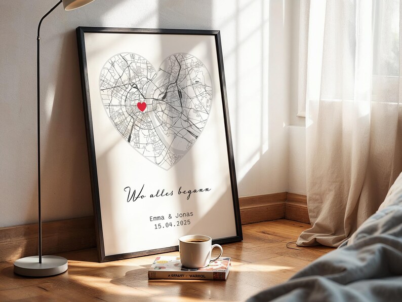 May include: Framed art print featuring a heart-shaped map with a red marker. The print includes the text "Wo alles begann", the names Emma & Jonas, and the date 15.04.2025. A coffee cup and a book are in the foreground.
