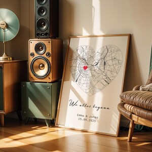 May include: A framed map print in the shape of a heart, with a red heart marking a location. The print includes the text "Wo alles begann" and the names Emma & Jonas with a date. The print is leaning against a wall.