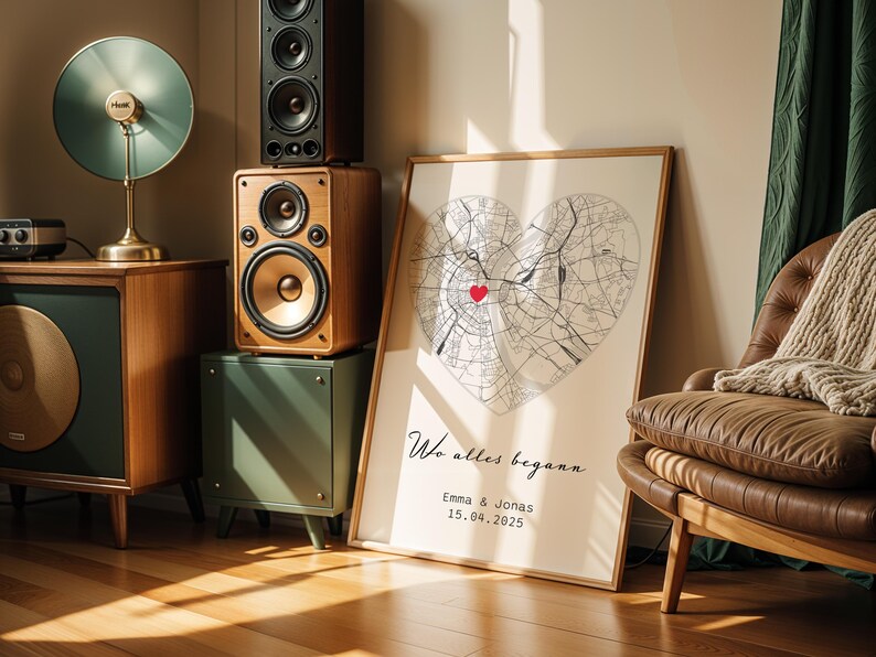 May include: A framed map print in the shape of a heart, with a red heart marking a location. The print includes the text "Wo alles begann" and the names Emma & Jonas with a date. The print is leaning against a wall.
