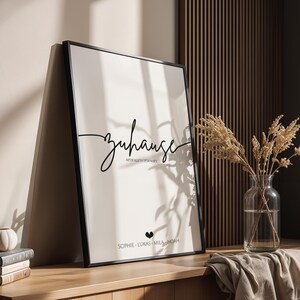 May include: A framed print with the word "Zuhause" in elegant script. Below the word are additional lines of text and names. The print is in a black frame and leans against a wall. A vase of dried flowers and a stack of books are nearby.