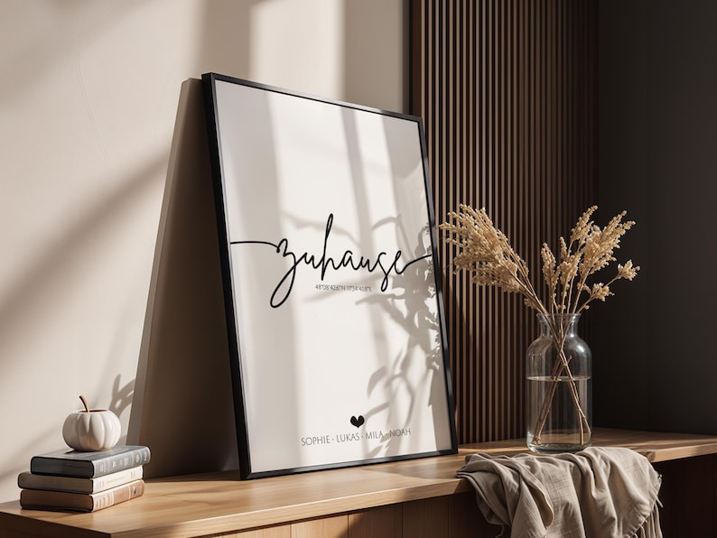 May include: A framed print with the word "Zuhause" in elegant script. Below the word are additional lines of text and names. The print is in a black frame and leans against a wall. A vase of dried flowers and a stack of books are nearby.