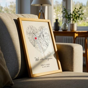 May include: A framed print featuring a heart-shaped map with a red marker. The print includes the text "Wo alles begann" and the names "Emma & Jonas" with a date. The frame is light-colored, and the print is displayed on a gray sofa.