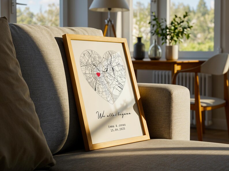 May include: A framed print featuring a heart-shaped map with a red marker. The print includes the text "Wo alles begann" and the names "Emma & Jonas" with a date. The frame is light-colored, and the print is displayed on a gray sofa.