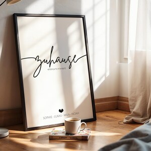 May include: A framed art print with the word "Zuhause" in elegant script, coordinates, and names. A coffee cup sits on a book in front of the print. The print is in a black frame, leaning against a wall.