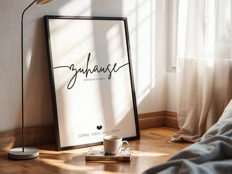 May include: A framed art print with the word "Zuhause" in elegant script, coordinates, and names. A coffee cup sits on a book in front of the print. The print is in a black frame, leaning against a wall.