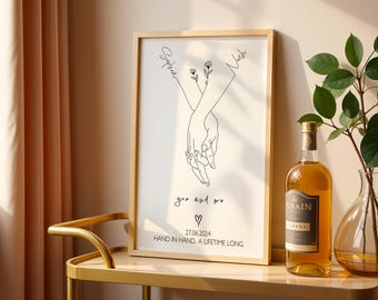 May include: A framed print with a minimalist line drawing of two hands intertwined, with the names "Sophia" and "Noah" above. The text "you and me", a heart, date, and the phrase "HAND IN HAND, A LIFETIME LONG" are also included.