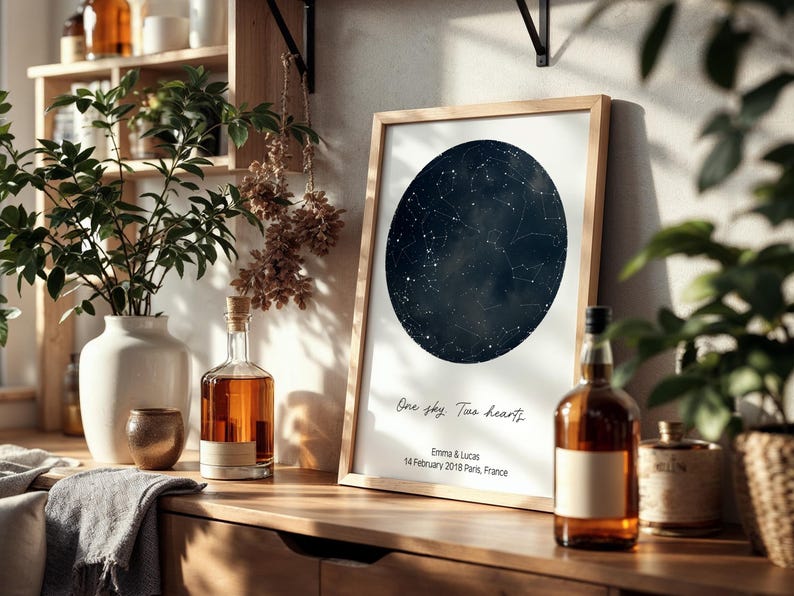 Peut inclure: Impression encadr&eacute;e d'une carte du ciel avec le texte "One sky, Two hearts" et les noms Emma & Lucas, dat&eacute;e du 14 f&eacute;vrier 2018, Paris, France. L'impression est sur une surface en bois avec des bouteilles et des plantes.