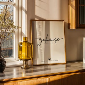 May include: A home decor scene featuring a framed print with the word "zuhause" in script, a yellow glass vase, a brown vase with dried flowers, and two cylindrical vases. The scene is set on a wooden cabinet near a window.