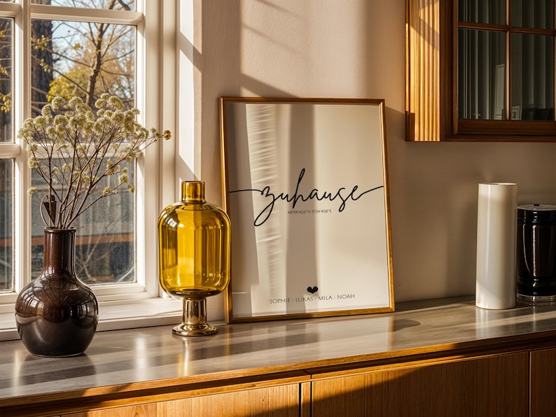 May include: A home decor scene featuring a framed print with the word "zuhause" in script, a yellow glass vase, a brown vase with dried flowers, and two cylindrical vases. The scene is set on a wooden cabinet near a window.