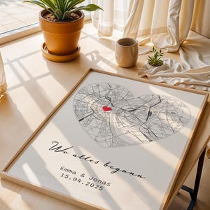 May include: A framed map print featuring two overlapping heart-shaped city maps in black and white, with a red heart marking a location. The print includes the text "Wo alles begann" and names and a date.